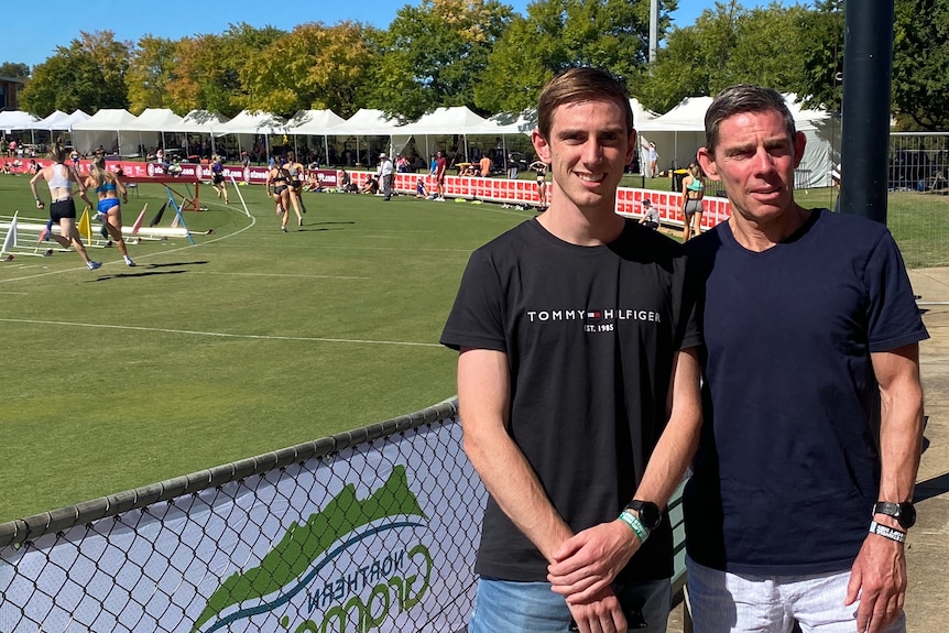 A young man and an older man stand beside an athletics track where women are running.