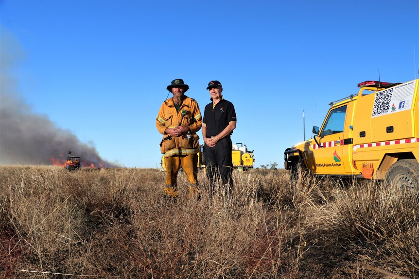 Two firefighters standing next to each other on a dry, grassy plain.