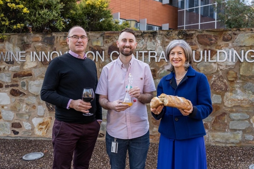 A man holding a wine glass, another man holding a beaker and a woman holding a loaf of bread