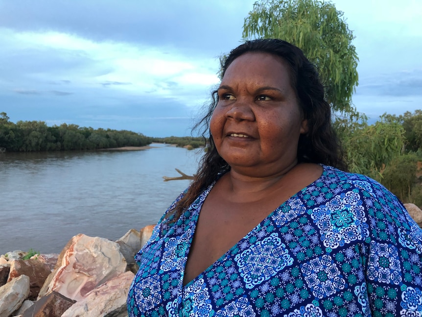 Image of an Aboriginal woman wearing a blue patterned top, standing on the banks of the Fitzroy River with a grey sky.