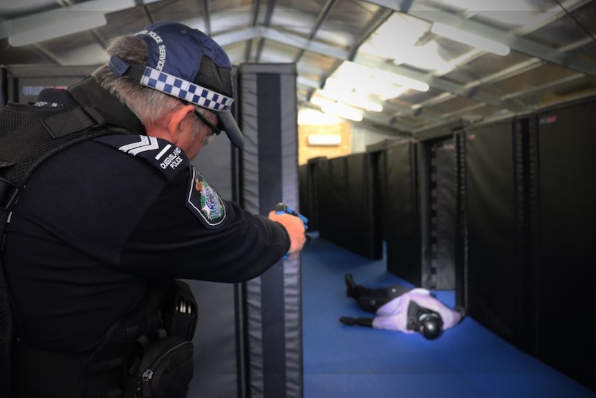 Over the shoulder photo of a police officer pointing a blue training pistol at a dummy on the ground during training.