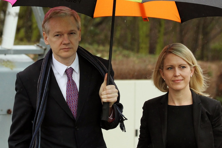 Julian Assange wears a suit holding an umbrella standing next to Jennifer Robinson