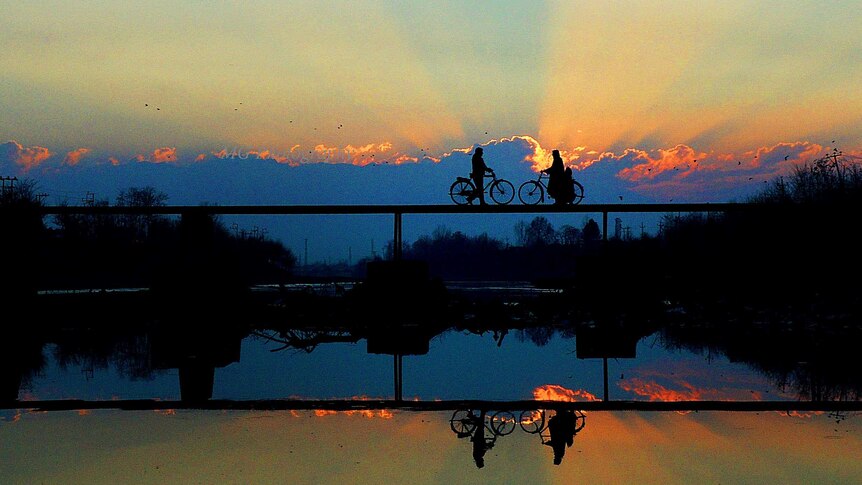 Two people riding bicycles across a bridge are silhouetted against the sunset and reflected in the still water of the river.