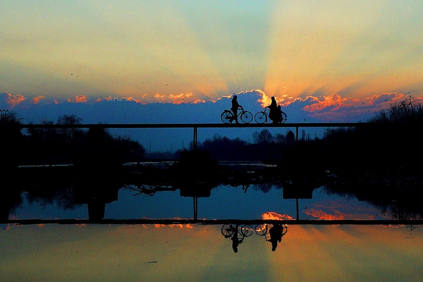 Two people riding bicycles across a bridge are silhouetted against the sunset and reflected in the still water of the river.