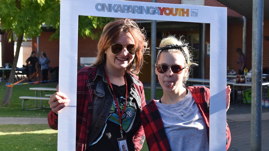 Two girls wearing aviator sunglasses smile at the camera as they hold up a white photo border around themselves.