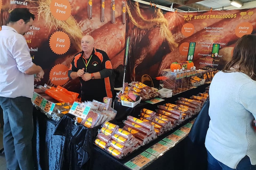 A bald man in an orange and black uniform speaks to a tall fellow from behind a meat market stall.