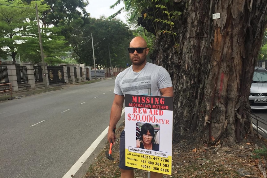 Man holding a sign for his missing mother standing on the side of a road.