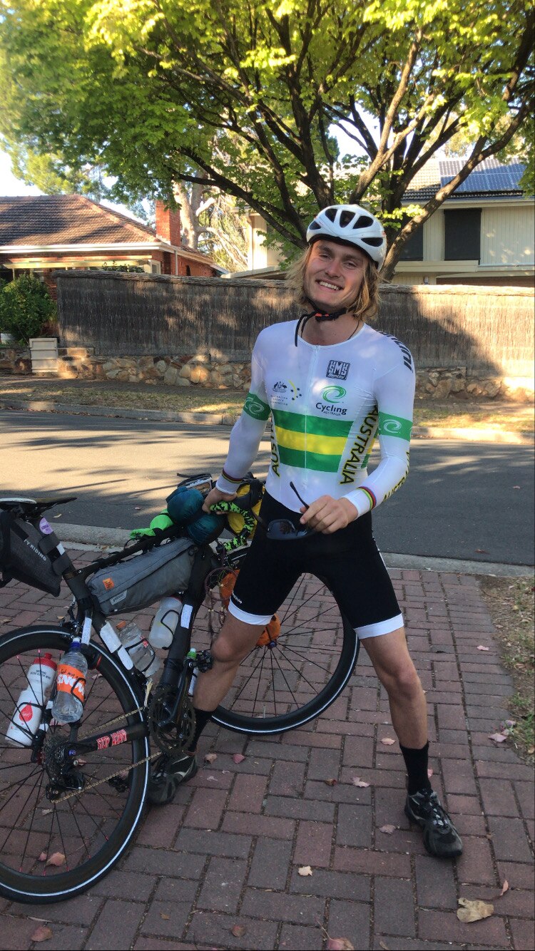 A young man with a big smile and wearing cycling gear stands with his bicyle