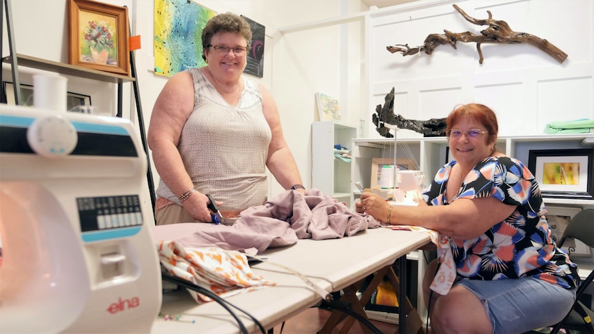 Two smiling women around a sewing table with fabric on it.