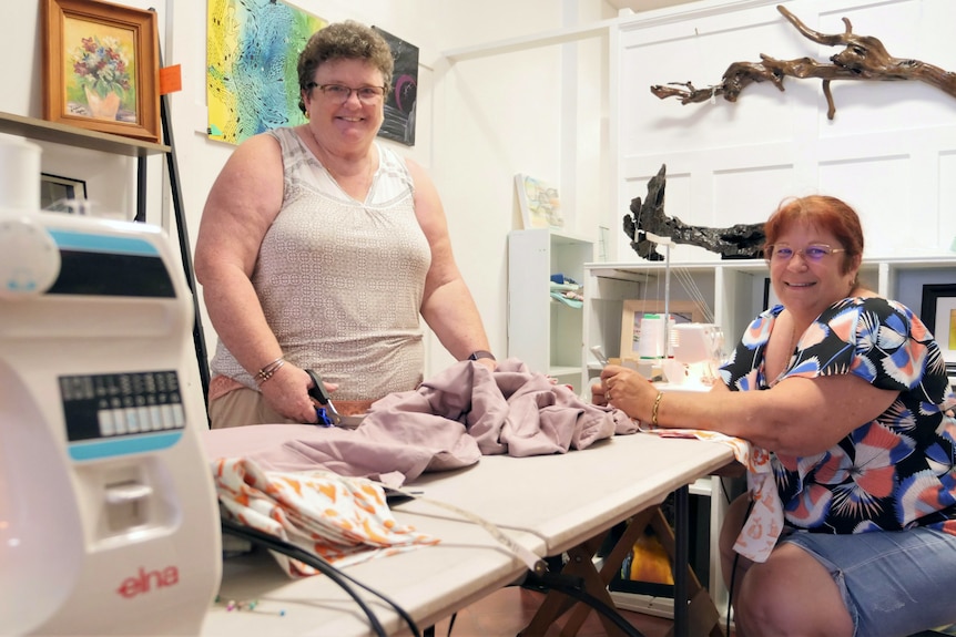 Two smiling women around a sewing table with fabric on it.