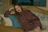 Emma Stone reclines on a plush mustard sofa, with her legs up as she rests a fist on her head and looks left. 