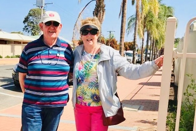 An older couple smiling and standing next to a fence, with palm trees near the road in the background