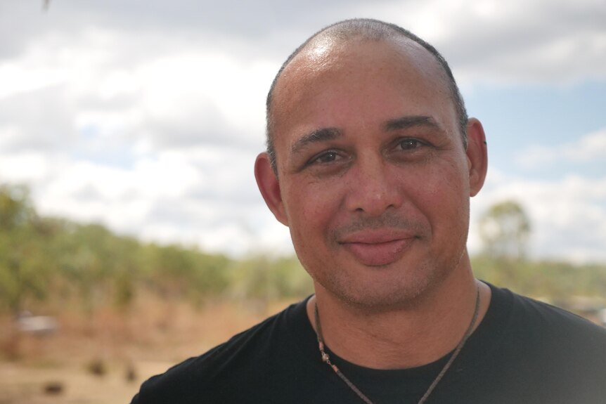 A man in a black T-shirt standing and smiling, in front of red dirt and scrub.