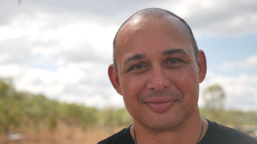 A man in a black T-shirt standing and smiling, in front of red dirt and scrub.