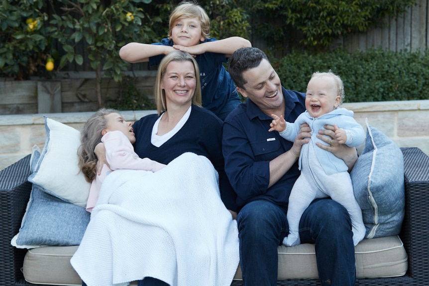 A family of five, including a girl who is being cradled by her mother, sit on an outdoor lounge