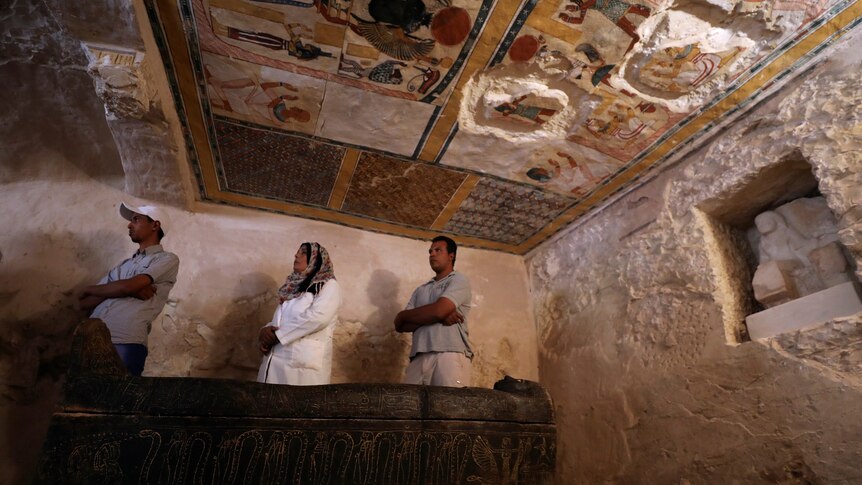 Two men and a woman wearing a headscarf stand in the tomb, beneath ad colourful painted ceiling showing Egyptian figures