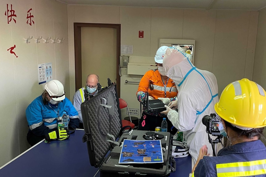 Five people in a cargo ship room, large black suitcase-style box, two people fiddling with Wi-Fi modem.