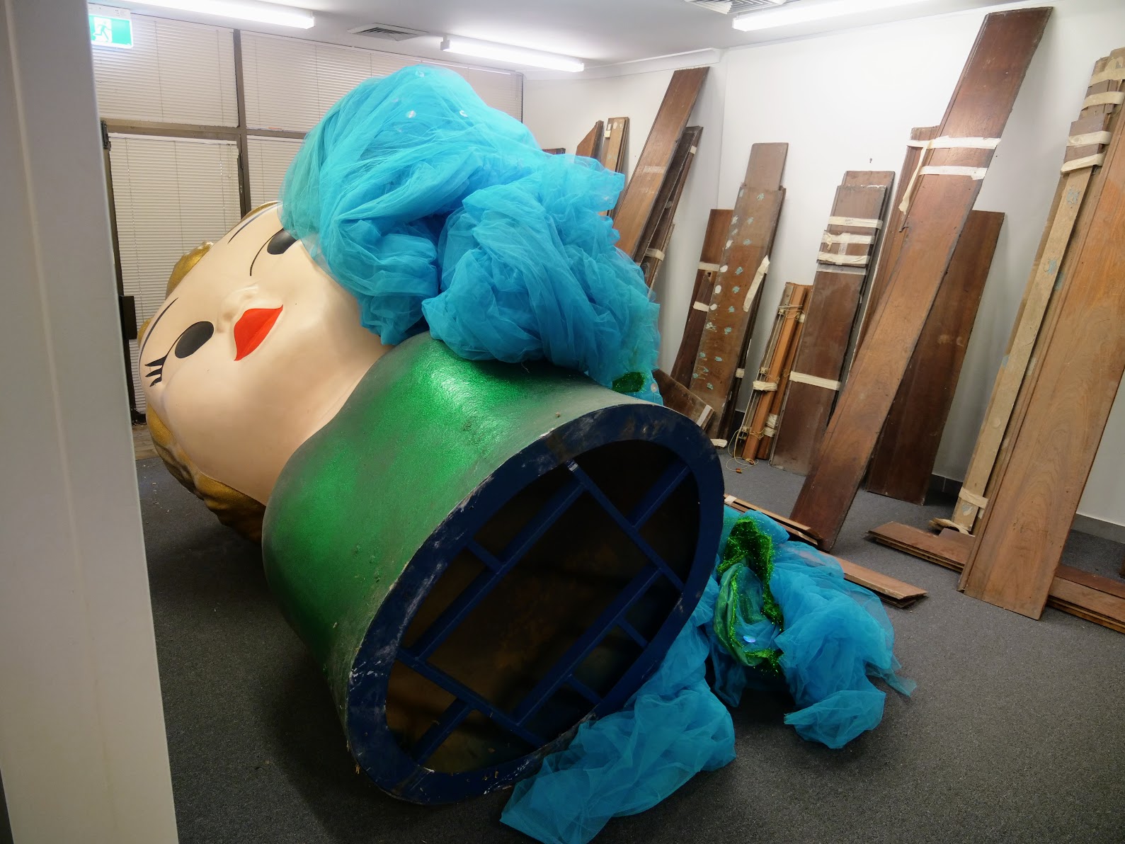 Uma boneca grande com batom vermelho e corpo verde está sob o tule azul guardado