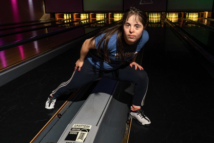 A woman steps dramatically over the gutters of two lanes in a darkened bowling alley.