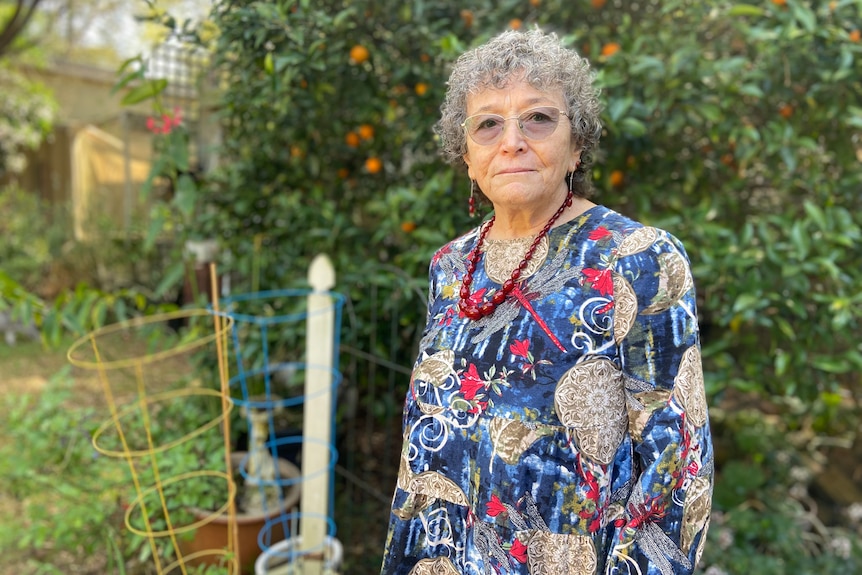 an older woman wearing glasses standing in front of a garden