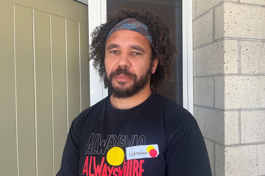 A man with dark curly hair and a beard, wearing a bandanna and a shirt with an Indigenous slogan on it.