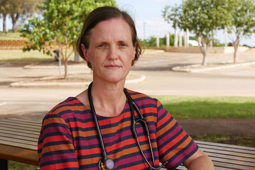 A woman wearing a stethoscope around her neck, sitting in a park with a serious expression on her face.