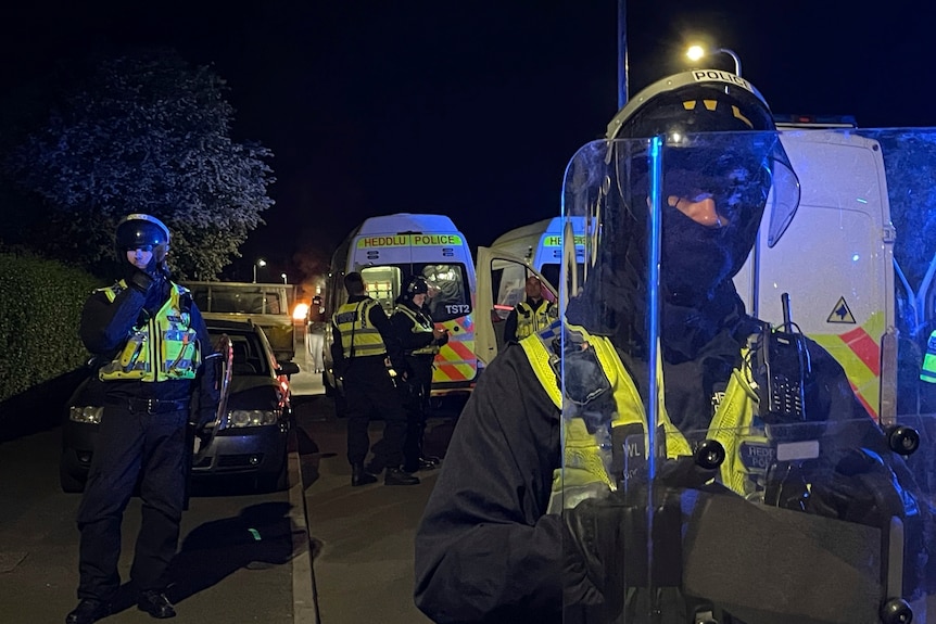 a police officer in riot gear stands at forefront with other officers and a police van behind