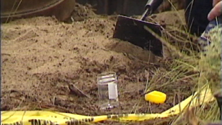 TV still of a man's hand near dirt with a small plastic container, a yellow lid and evidence tape in the foreground.