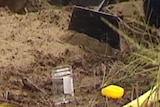TV still of a man's hand near dirt with a small plastic container, a yellow lid and evidence tape in the foreground.