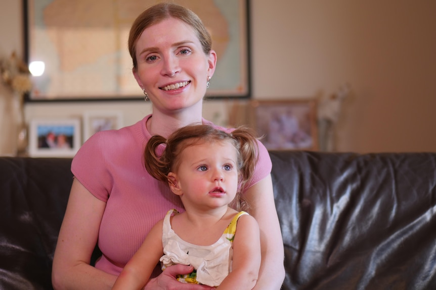 A woman sits on a couch and smiles, with a toddler sitting in her lap.