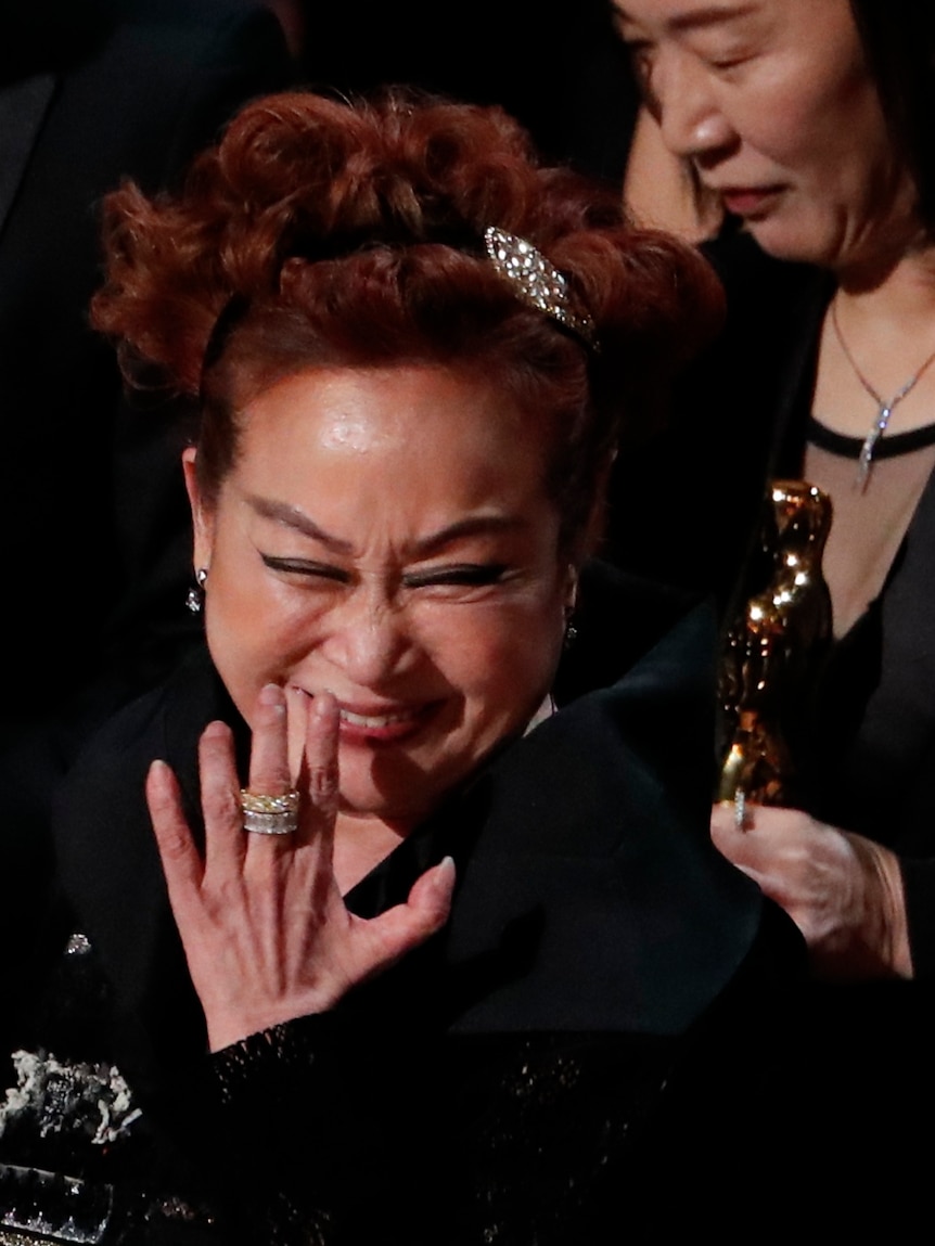 A woman with curled red hair laughs and puts her hand to her mouth with someone holding a gold Oscar statue behind her.