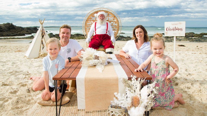 A boy and a girl sit with their parents at a low wooden table on a beach, with Santa sitting in a large chair behind them.