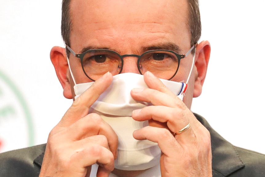 You view a balding man with thin wire glasses up close wearing a white face mask with the French tricolour in its top-right.