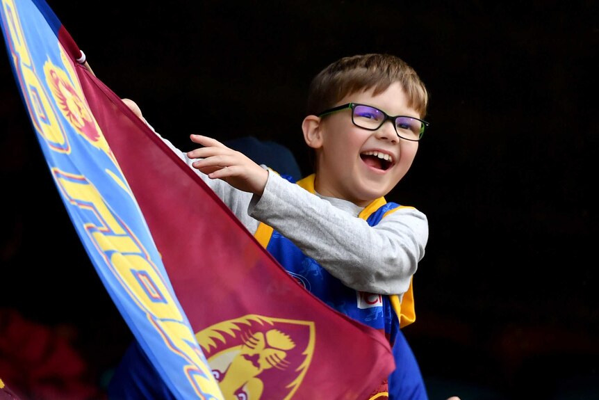 A young Brisbane Lions fan waves a flag as he smiles in the stands at the Gabba.