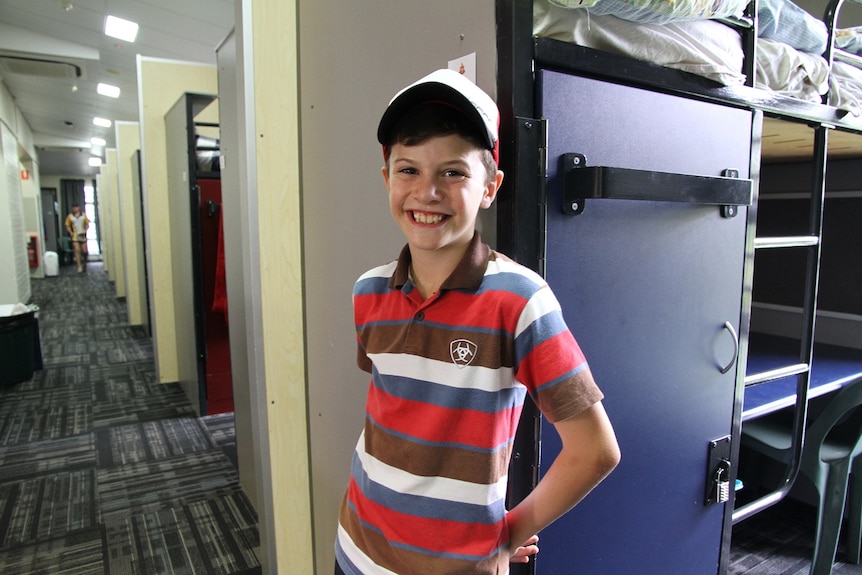 A boy wearing a cap and a striped short standing near is bunk bed at boarding school.