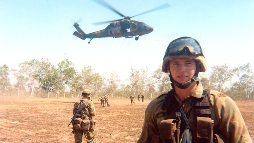 An Australian soldier standing with a helicopter hovering in the background, and six other soldiers in the distance.