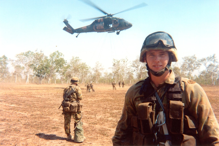 An Australian soldier standing with a helicopter hovering in the background, and six other soldiers in the distance.