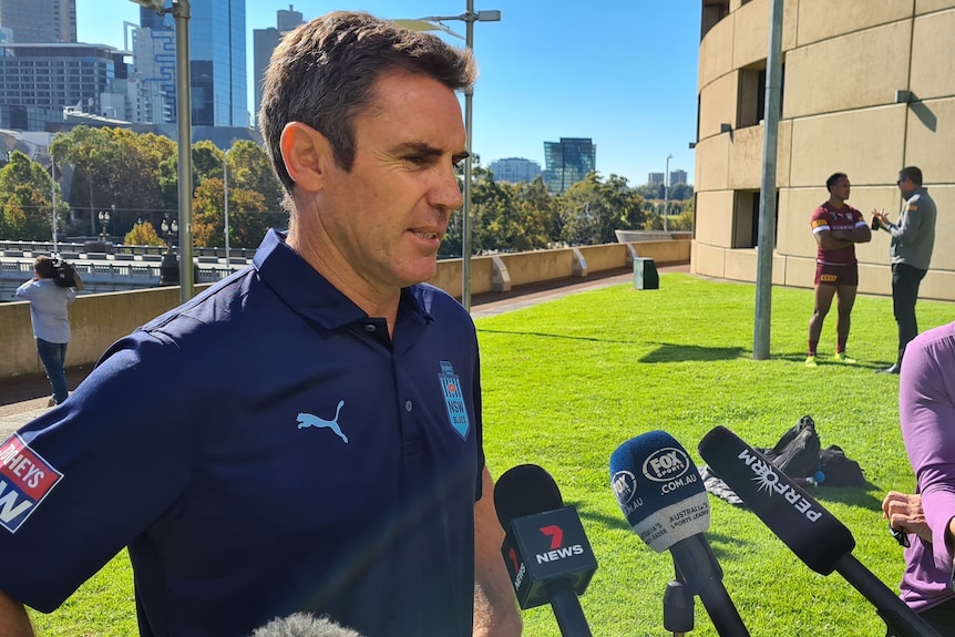 A man in a media scrum outside on a sunny day.