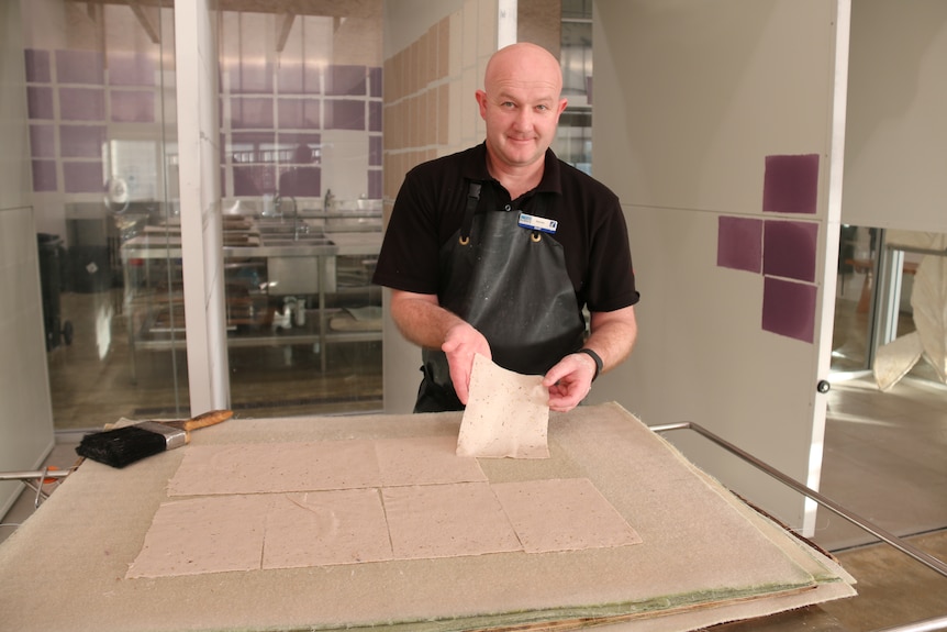 Man wearing apron arranges wet handmade paper on a table.