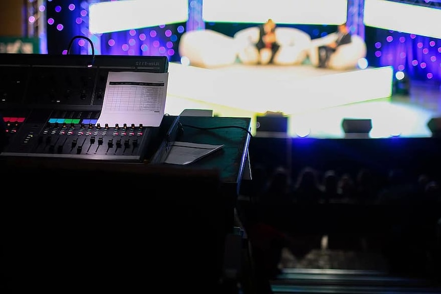 A control board in front of a television studio with two people on a stage.