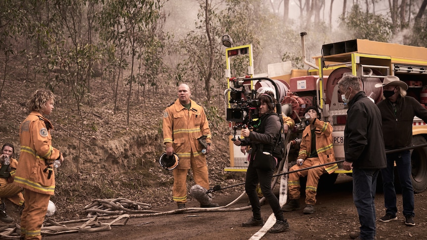 Actors wearing fire gear standing on road near fire truck while camera crew films.