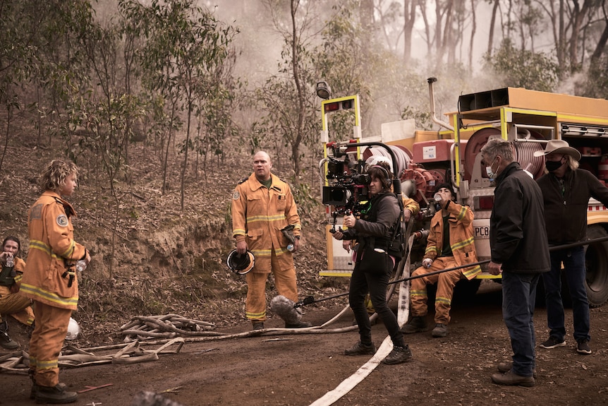 Actors wearing firefighters' clothes standing on the road near a fire truck while the film crew films.