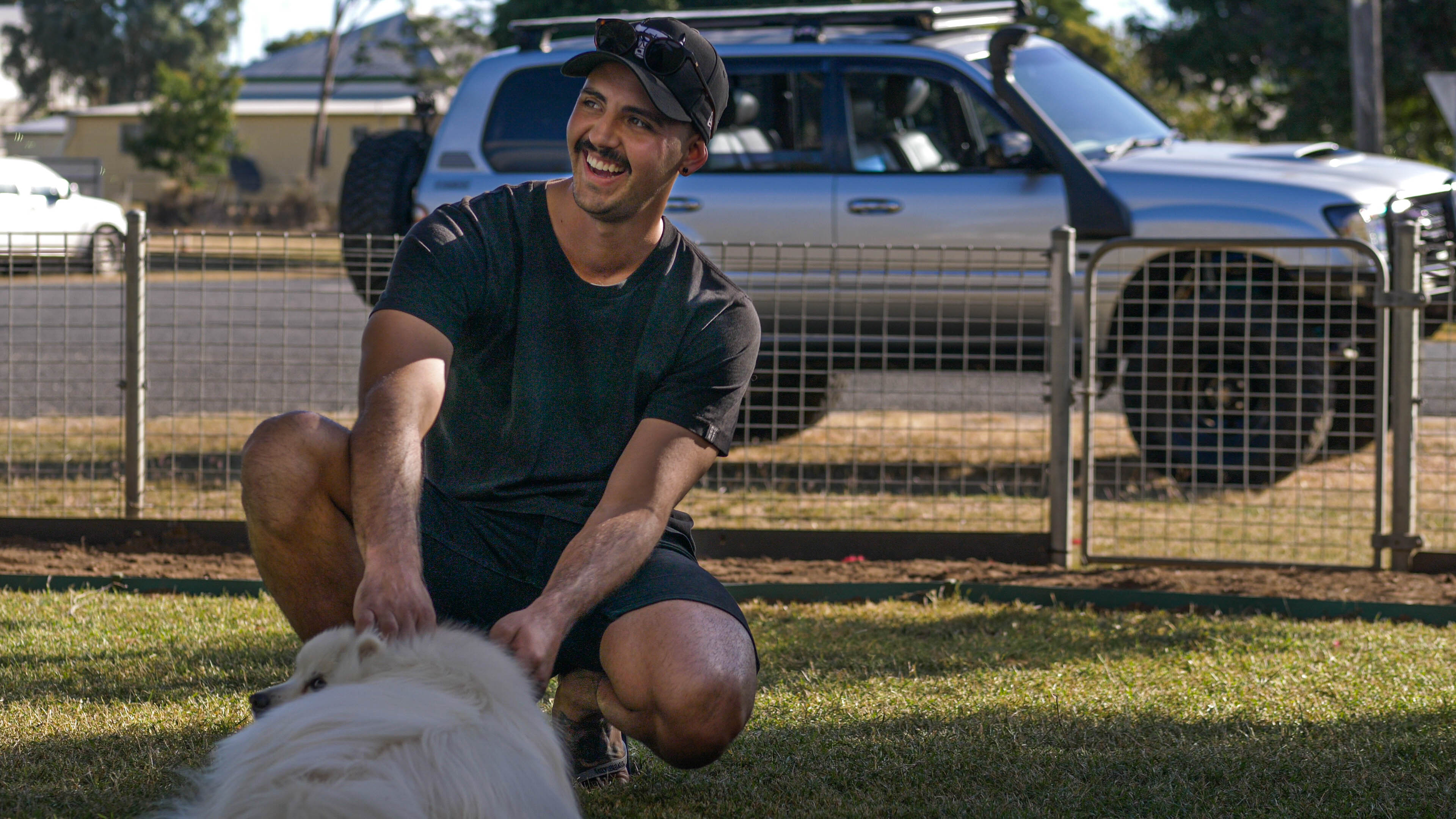 A man wearing a black t-shirt, cap pats a dog, his diesel 4WD is in the background.