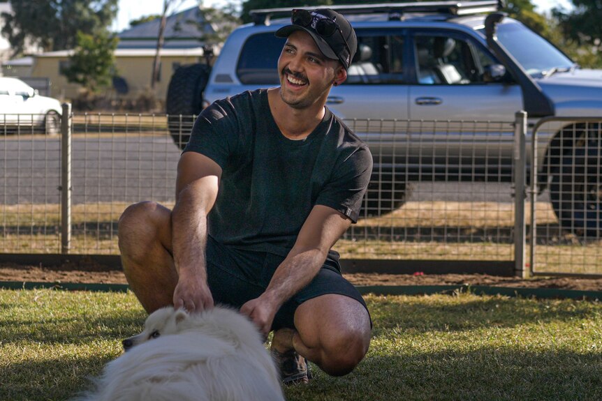 A man wearing a black t-shirt, cap pats a dog, his diesel 4WD is in the background.