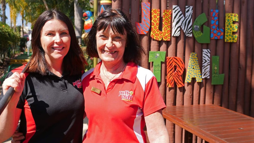 Two women stand in front of a putt putt course with a colourful sign that says jungle trail.