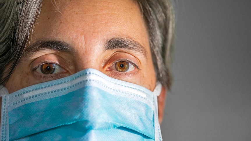 Close up picture of a grey haired woman wearing a surgical mask. Her brown eyes stare directly at the camera.
