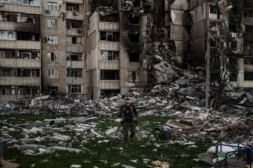 A man in military uniform walks through rubble outside a badly damaged residential building.