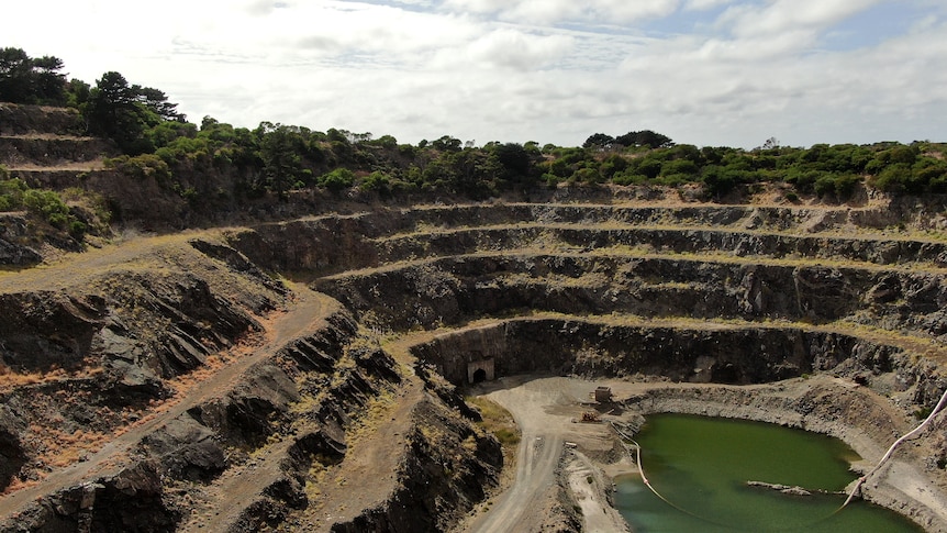 A large, multi-layered open pit mine, which is overgrown. Green water sits in the pit's lowest level.