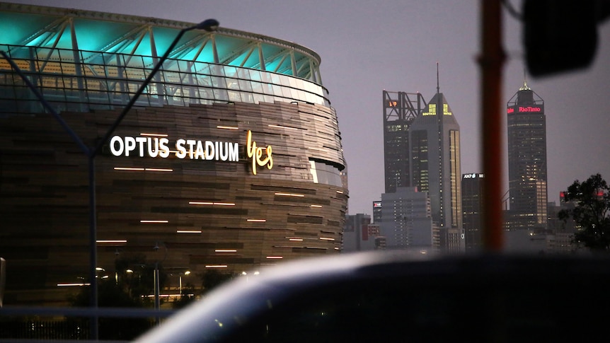 Perth Stadium at night with the CBD skyline behind it a car passes, "Optus Stadium Yes" is on the side of the building