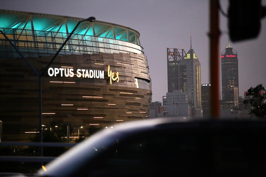 Perth Stadium at night with the CBD skyline behind it a car passes, "Optus Stadium Yes" is on the side of the building